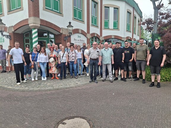 Eine große Gruppe von Menschen steht dicht nebeneinander vor einem Gebäude mit grün-weißen Fenstern und einer Aufschrift „Potts Brau- und Backhaus“.