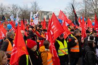 Das Foto zeigt eine große Menschenmenge bei einer Demonstration. Die Teilnehmenden tragen viele rote Fahnen mit weißer Aufschrift, auf denen Eisenbahn- und Verkehrsgewerkschaft (EVG) steht. Viele Personen tragen Warnwesten in Gelb oder Orange. Einige halten auch andere bunte Schilder und Fahnen in der Hand, z. B. mit Symbolen oder Zeichnungen. Im Hintergrund sind winterliche, kahle Bäume sowie ein modernes Regierungsgebäude zu sehen – es handelt sich um eine Demonstration in Berlin. Die Stimmung wirkt engagiert und friedlich. Es ist Tag, das Wetter scheint kühl, aber trocken zu sein.