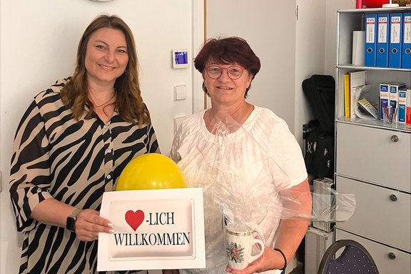 Zwei Personen stehen in einem Büro nebeneinander und halten ein Geschenk sowie ein Schild. Die beiden Personen stehen vor einer weißen Wand. Eine Person hält einen gelben Ballon und ein gerahmtes Schild mit dem Text „Herzlich Willkommen“, bei dem ein rotes Herz das Wort ergänzt. Die andere Person hält eine Tasse, die in durchsichtige Folie eingepackt ist. Beide schauen in Richtung Kamera. Rechts im Bild steht ein Regal mit Ordnern, Broschüren und einem Telefonkoffer oder einer Tasche. Hinter ihnen ist ein Lichtschalter und ein kleines Gerät an der Wand angebracht.